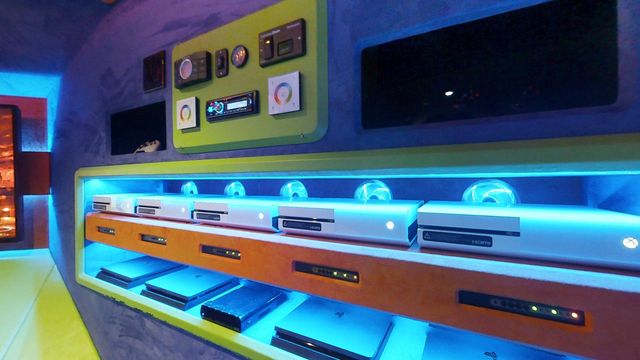 The AV rack compartment: a row of gaming consoles on a lit shelf beneath the lighting, audio and generator controls