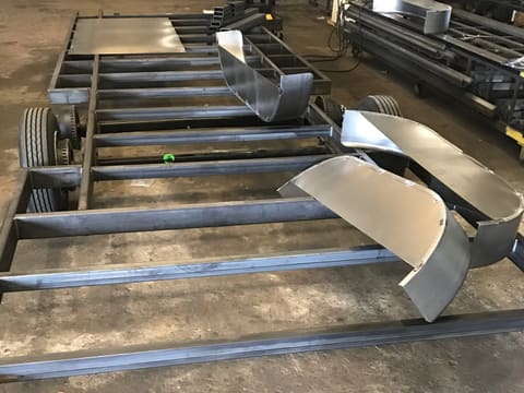 A bare welded steel trailer chassis on its tandem axles on the shop floor, with steel fenders resting on the crossmembers