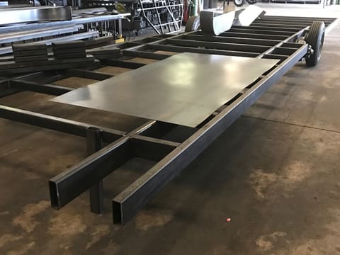 A bare welded steel trailer chassis on the shop floor, with a loose steel floor sheet laid across the crossmembers and a wheel at the far end
