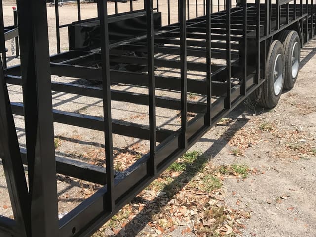 A bare welded black steel trailer floor frame on tandem axles, standing on gravel, before any floor, walls or skin go on