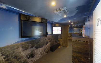 Inside a traveling museum build: a large screen framed by speakers on the left, a desert landscape mural wrapping the walls and ceiling, and a display panel on the right