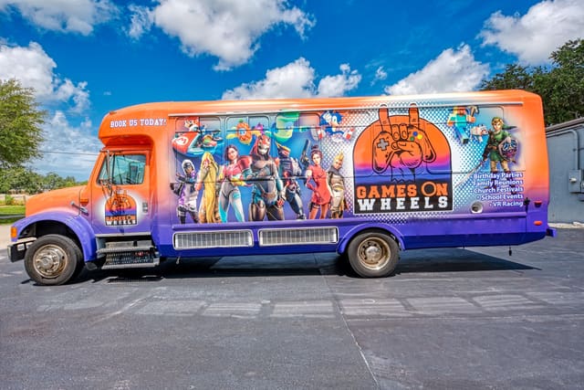 A bus converted into a gaming bus, seen from the side: a full custom wrap along the body in the owner's branding, with game artwork across the panels and the entrance door at the front