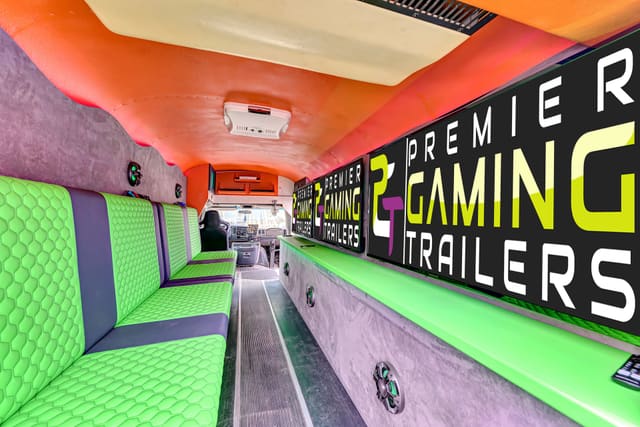 Inside the second converted gaming bus: green hex-stitched benches down one side, screens showing the PGT logo, an orange ceiling and the driver's seat and dashboard visible at the far end