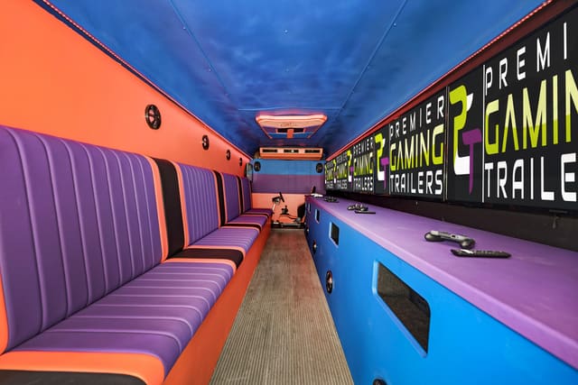 Inside a converted gaming bus: screens along one wall showing the PGT logo, purple channel-stitched benches facing them, a blue ceiling and a racing seat at the far end