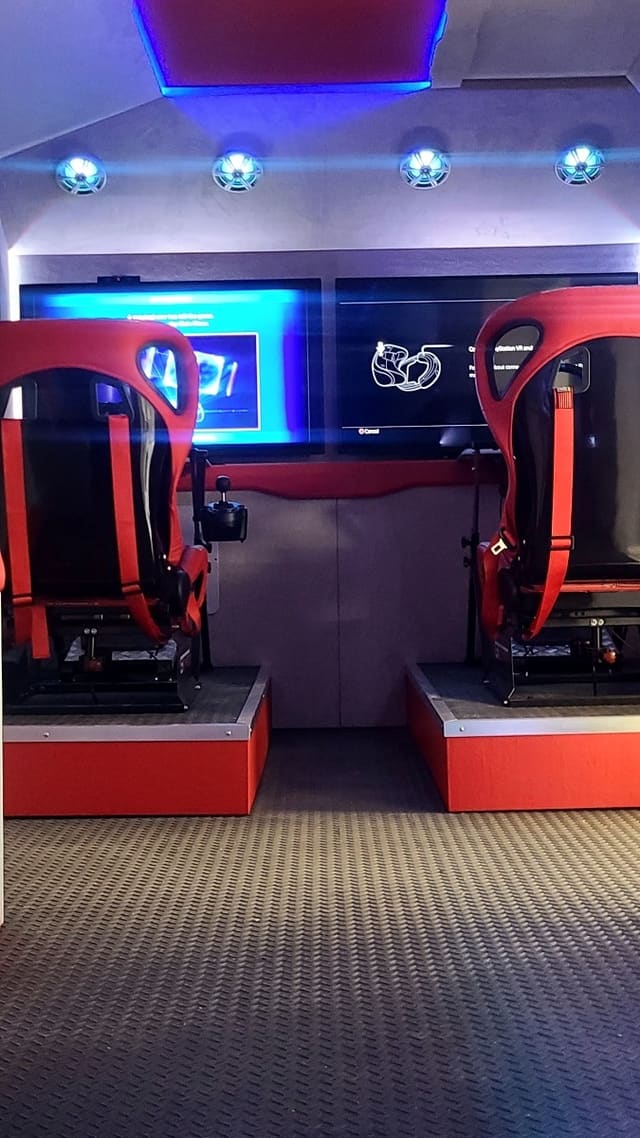 Two racing simulator stations side by side in a finished build: molded racing seats on raised platforms, a joystick and pedals at each one, and a screen on the wall in front of them