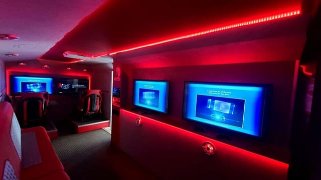 Inside a custom racing simulator trailer: two racing seats on platforms with wheels and pedals, screens on the wall beside them and red LED light along the ceiling