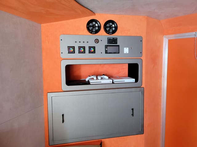 The cabinet at the front of a Gamers Paradise: two speakers, a panel of lighting controllers and a radio, and a shelf holding consoles and controllers
