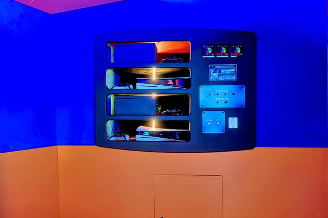 The console cabinet at the front of a Gamers Paradise: four lit shelves of consoles, a stereo, lighting controllers and labeled switch panels set into a blue wall above an orange base