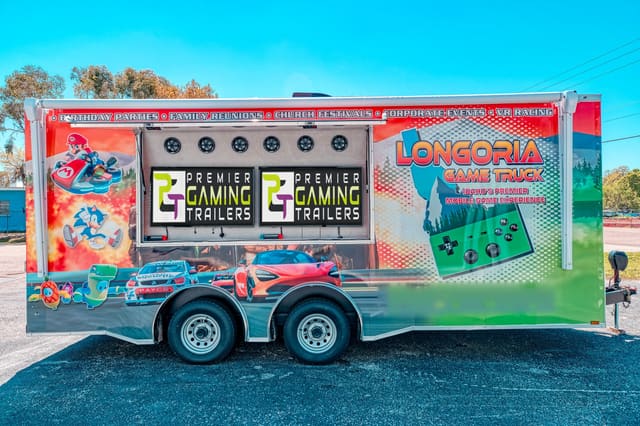A Gamers Paradise from the side with its exterior screen hatch raised, two screens under the hatch showing the PGT logo, a full custom wrap along the body and tandem axles beneath