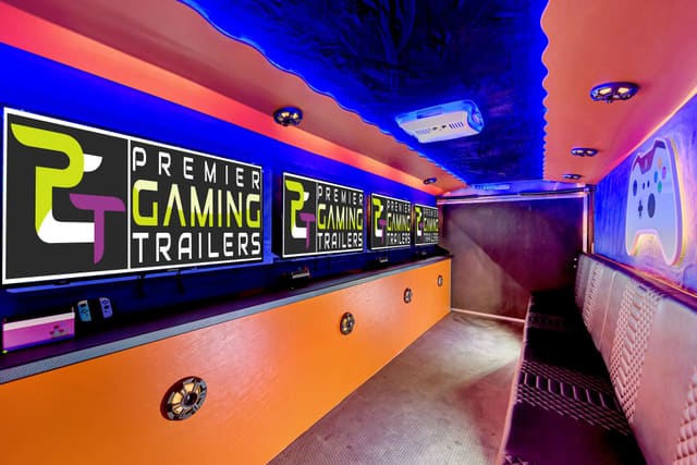 Inside a Gamers Paradise: PGT screens along an orange counter on the left, a blue LED ceiling, and a lit game controller shape on the end wall beside a black and white bench
