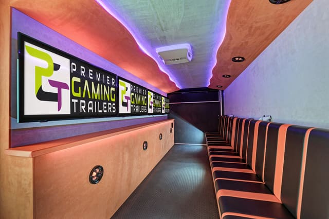Inside a Gamers Paradise: four screens along the wall showing the PGT logo, an orange counter beneath them, striped benches facing the screens and LED light along the ceiling