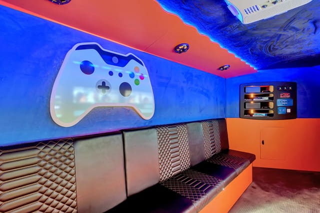 A corner of a Gamers Paradise: a large white game controller shape lit on a blue wall under an orange ceiling with blue LED, black diamond-stitched benches below it and a console cabinet in the corner