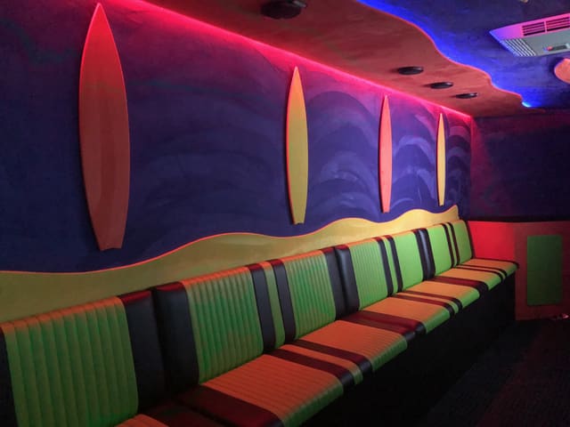 Inside a Gamers Paradise: surfboard shapes mounted on a blue and purple wall above a bench with green, orange and black stripes, with red and magenta LED light along the edge of the ceiling