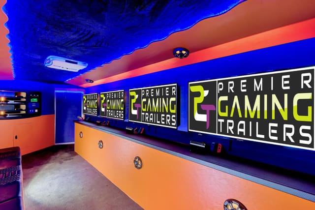 Inside a Gamers Paradise: three PGT screens on the wall above an orange counter with consoles and controllers on it, and a blue ceiling edged with LED light