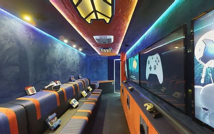 Inside a Gamers Paradise owner build: orange and navy benches down the left under a lit ceiling panel, facing a row of screens with controllers on the counter opposite