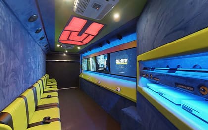 Inside a Gamers Paradise owner build: yellow benches along the left, a red lit ceiling feature overhead, and screens above a blue-lit shelf of consoles on the right