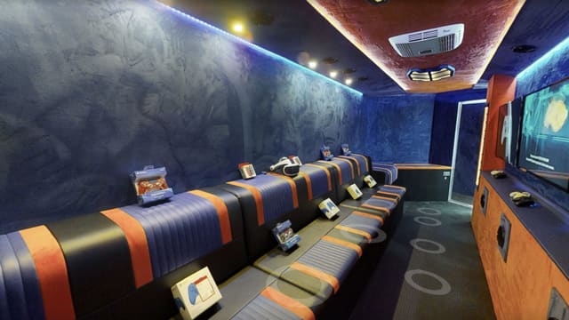 Opening view of the Gamers Paradise owner build tour: navy and orange striped bench seating with boxed controllers and a VR headset laid out, blue suede walls, and screens on the orange wall at the far end
