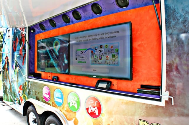 An exterior screen mounted in its open hatch on the side of a gaming theater, switched on and showing its menu, with speakers in a row above it and console logos on the wrap below