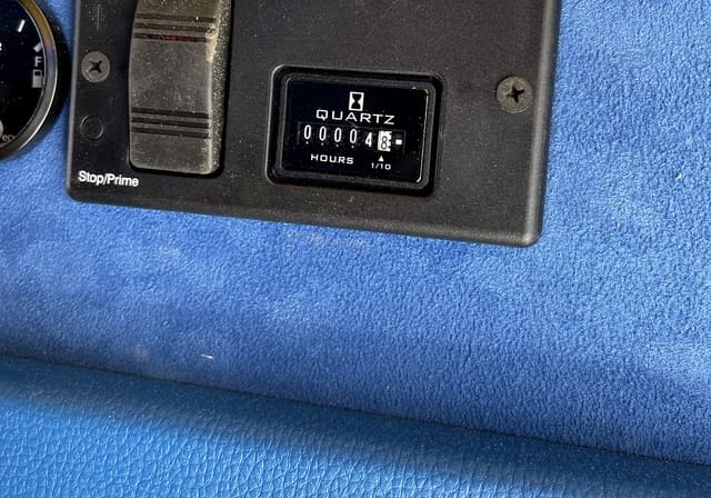 The control panel of an onboard generator in a finished trailer: a fuel gauge, a start and stop switch marked Stop and Prime, and an hour meter reading in tenths of an hour, set into a blue suede wall