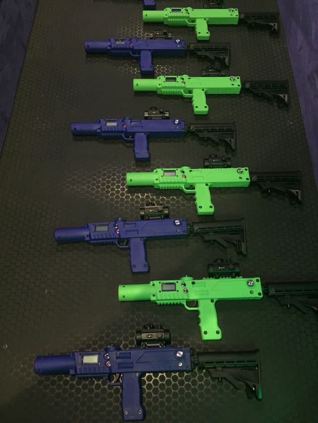 Laser tag blasters laid out in a row on a mat: green and blue units with small display panels, red indicator lights and sights fitted to the top rails