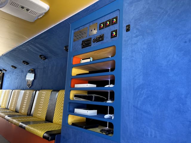 The console rack in a Gamers Utopia: consoles on lit shelves in a blue cabinet with controllers beside them, the lighting controllers and radio above, and quilted benches alongside