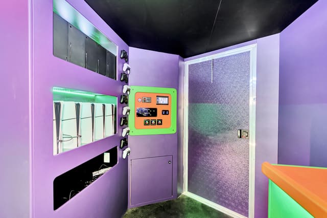 The front corner of a Gamers Utopia: a purple wall with controllers hung on hooks, lit shelves holding consoles, and an orange and green control panel with lighting dials beside a door