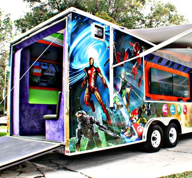 An owner's gaming theater set up outdoors with the side awning out, the exterior screen hatch open beneath it and the rear ramp down, wrapped in superhero and game artwork