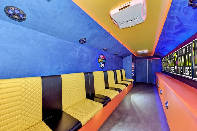Looking down the length of a Gamers Utopia: yellow diamond-quilted benches along the left, blue suede walls, PGT screens above an orange base on the right, and a blue and orange ceiling edged with LED light
