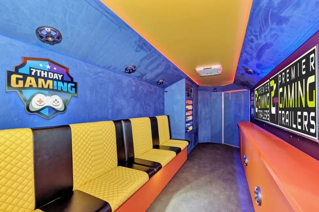 Inside a Gamers Utopia: yellow diamond-quilted benches on the left under a blue suede wall with an owner's logo, a console shelf at the far end, and an orange counter with PGT screens on the right