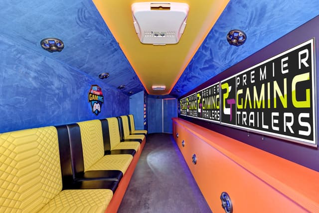 Inside a Gamers Utopia: screens along the wall showing the PGT logo, yellow diamond-quilted benches beneath them, a blue ceiling and the owner's logo panel at the far end