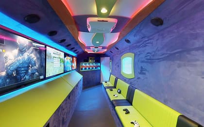 Inside a Gamers Utopia owner build: screens running down the left wall, lime benches opposite, and a pink and blue lit ceiling above a porthole window