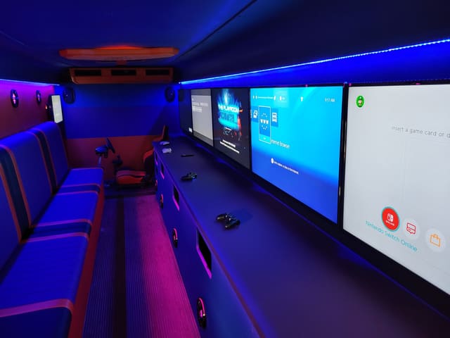 Inside a gaming bus conversion: a row of screens along the wall under a blue LED ceiling strip, blue and red benches facing them, and a racing seat with a wheel at the far end