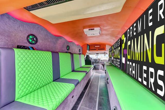 Looking down a converted gaming bus: lime green hex-stitched benches on the left, a PGT screen wall above a lime counter on the right, and an orange ceiling with a pink glow