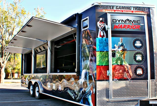 An owner's gaming trailer seen from the rear corner: the side awning raised on its struts, the exterior TV hatch open beneath it, the side door standing open, a full custom wrap across the body and rear doors, and tandem axles under the near side