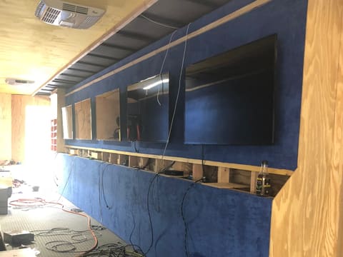 Fit out in progress inside a gaming trailer: a blue suede-feel wall going up, flat screens mounted in their bays, and cabling still loose on the floor