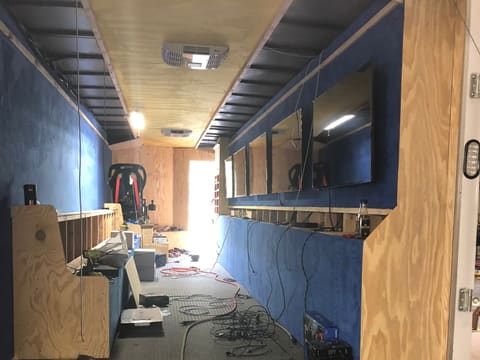 Looking in through the rear door of a gaming trailer mid fit out: screens mounted along a blue suede-feel wall, the bench framed in plywood, cabling on the floor