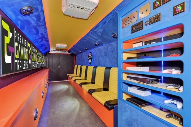 Inside a gaming trailer: blue suede walls, an orange counter under a row of wall-mounted screens, a yellow and black quilted bench, and a blue console rack holding PlayStation and Xbox consoles and controllers