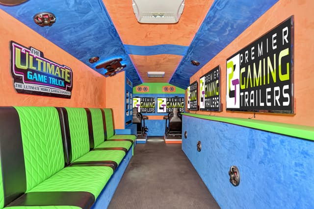 Lime benches with black stripes down the left side of a gaming trailer, three PGT screens on the right wall, an orange ceiling with blue bands, and two racing simulator seats at the far end