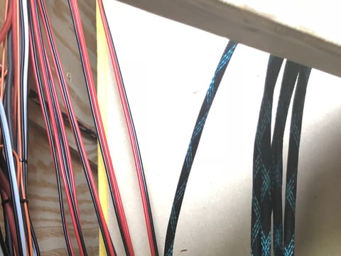 Red and black power cables and blue braided looms tied off down a plywood cabinet wall during the wiring stage of a gaming trailer build