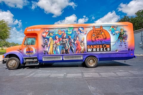 A bus converted into a gaming bus, seen from the side: a full custom wrap along the body in the owner's branding, with game artwork across the panels and the entrance door at the front