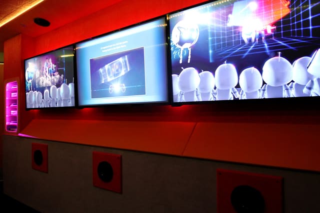 Screens in a row along the wall of a Mini Adventurer, with the lit console rack at the near end, speakers set into the bench front below and red and purple light across the ceiling