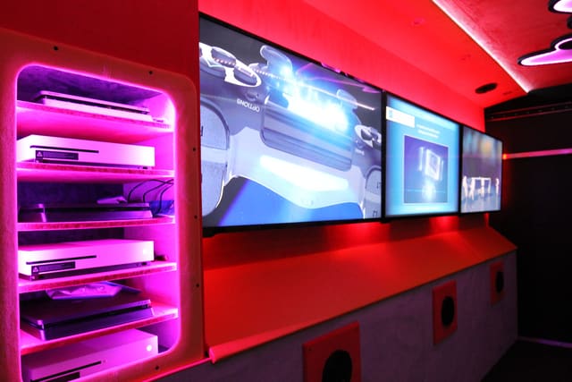 A console rack lit in magenta beside a row of three screens on the wall of a Mini Adventurer, with red LED light along the ceiling