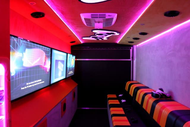 Inside a Mini Adventurer: three screens in a row along the wall, a striped bench beneath them with controllers and a VR headset on it, and a lit controller shape set into the ceiling