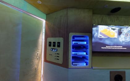 Inside a Mini Adventurer owner build: a wall panel of lighting controllers and a radio beside an open cubby holding consoles on blue-lit shelves, with a screen to the right