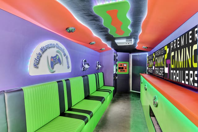 Interior of a finished gaming trailer: purple suede walls, an orange ceiling with a green wavy feature, a bright green and black bench, headsets on the wall, and a lit owner sign reading Filo's Xtreme Gaming Truck
