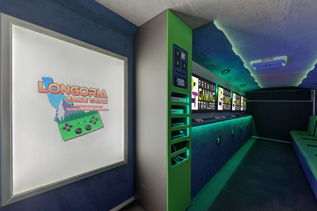 Interior of a finished gaming trailer: a lit lightbox sign reading Longoria Game Truck, a green console column with lit shelves of consoles, navy suede walls and a run of screens down the far wall