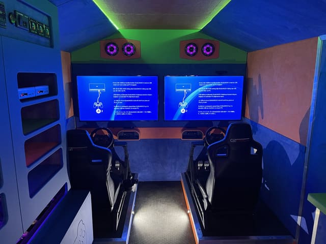 Two racing simulator stations side by side inside a trailer, each with a black racing seat, steering wheel and pedals facing its own screen, under a green LED ceiling with a pair of speakers over each screen