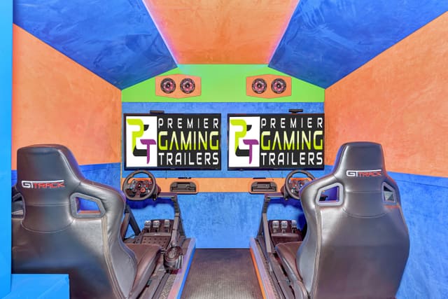 Two racing simulator stations inside a trailer: a GT Track seat, steering wheel, shifter and pedals at each one, facing its own screen, with speakers overhead and a blue and orange interior
