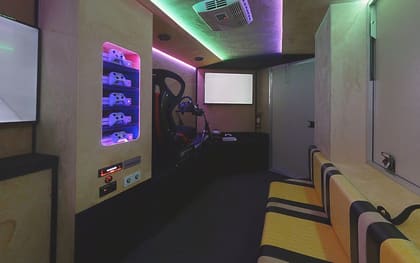 Inside a Virtual Reality owner build: controllers on purple-lit shelves beside a racing simulator rig, with a lit screen ahead and striped benches along the right